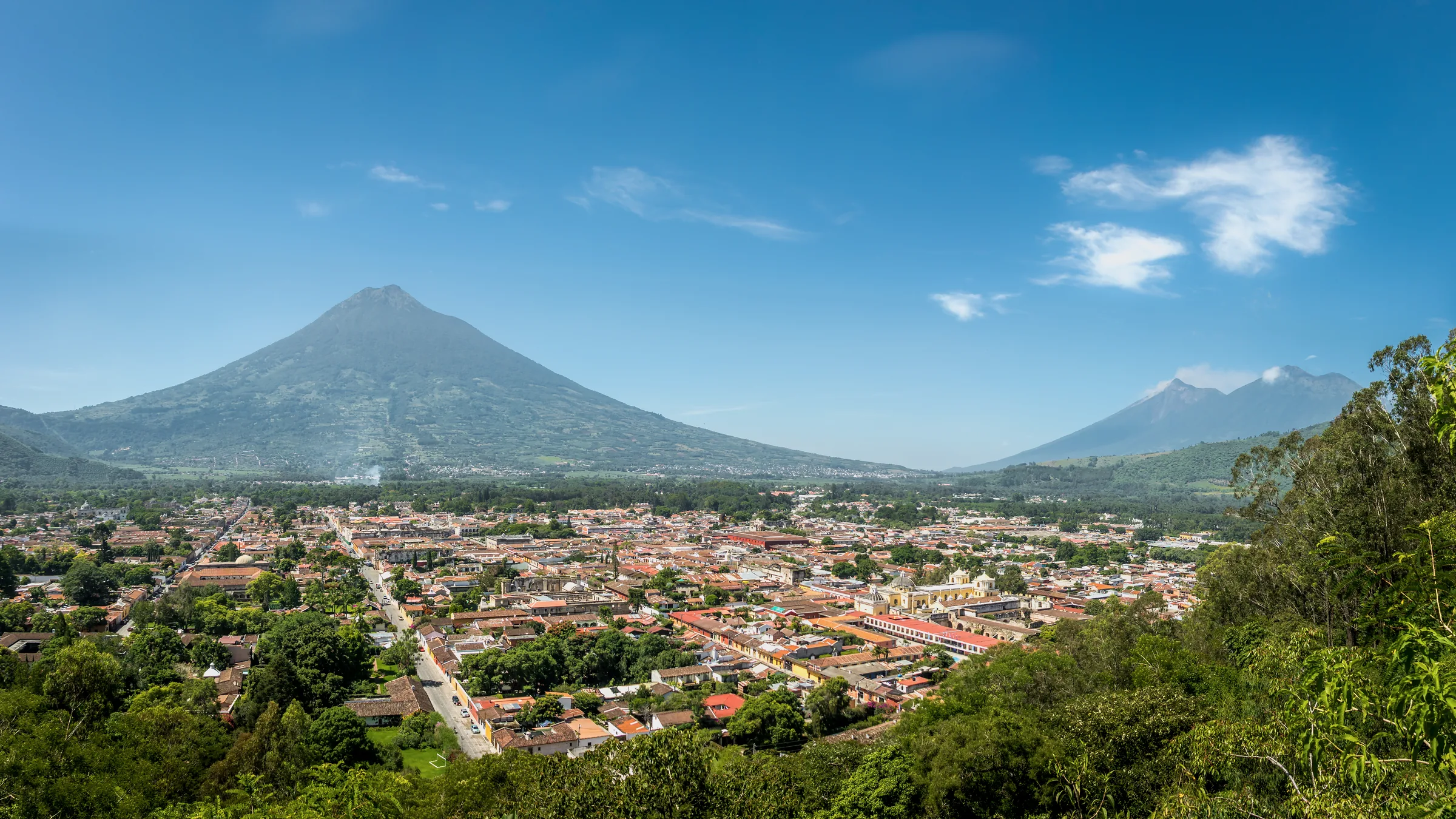Rooftops of Antigua Guatemala beneath a volcano, viewed from a vegetated hillside.