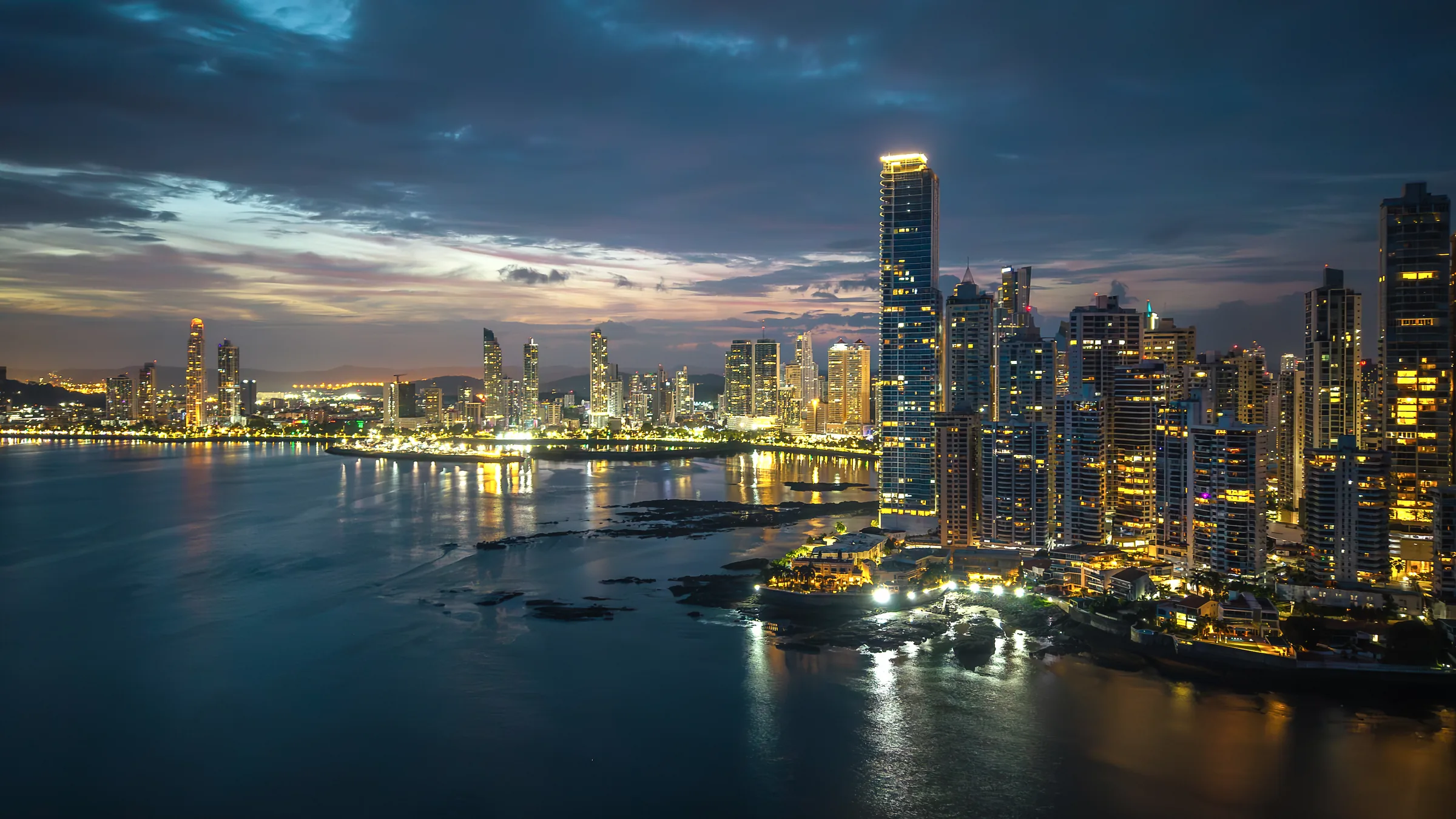 Illuminated skyscrapers beside Panama City Bay at dusk.