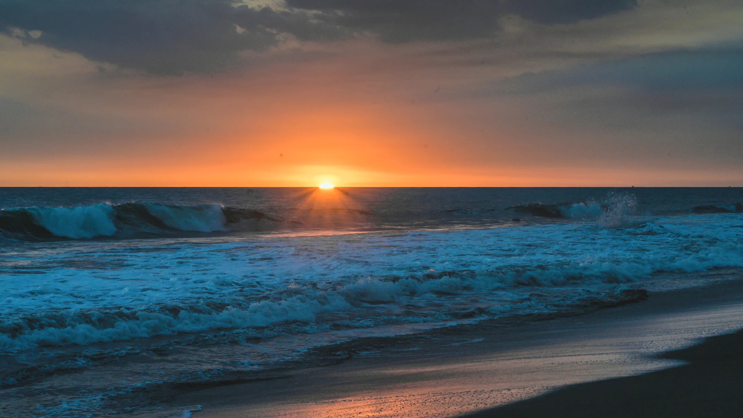 Waves reaching a dark beach as the sun meets the horizon.