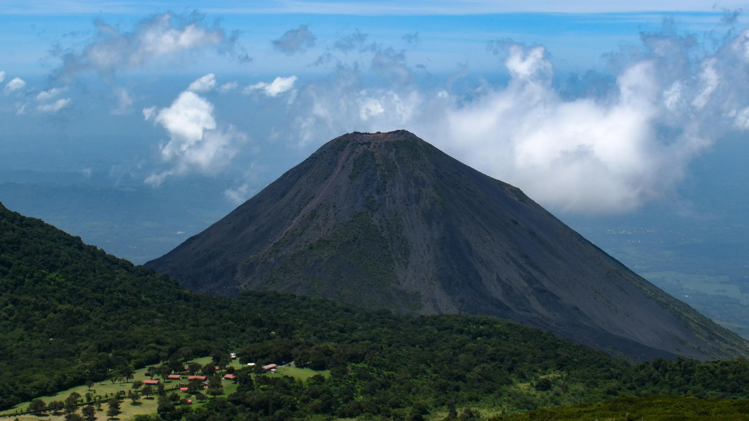 Volcanic cone above forested slopes beneath a cloudy sky.