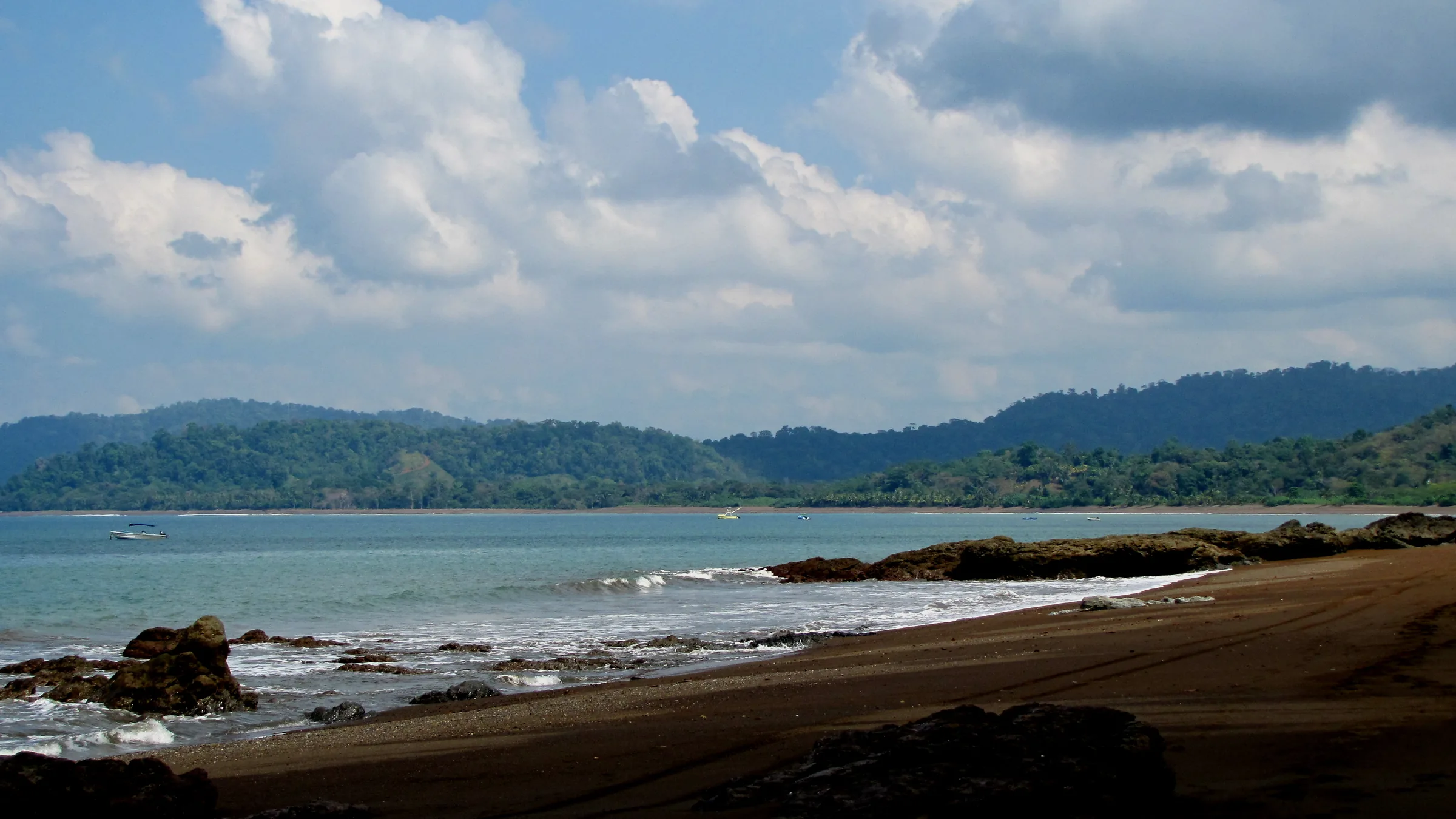 Dark sandy beach with waves and wooded hills across the bay.