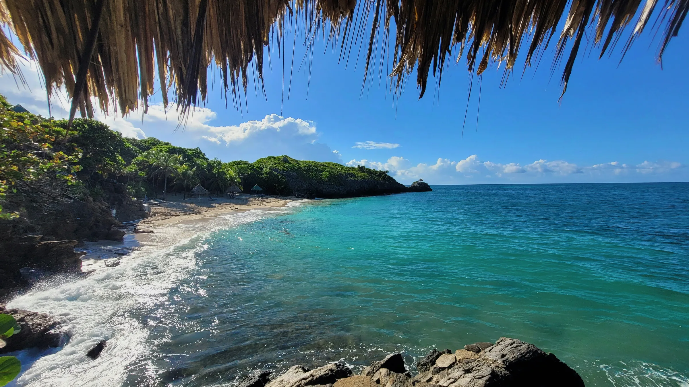 Beach and wooded coast of Roatán seen beneath a palm-thatch roof.