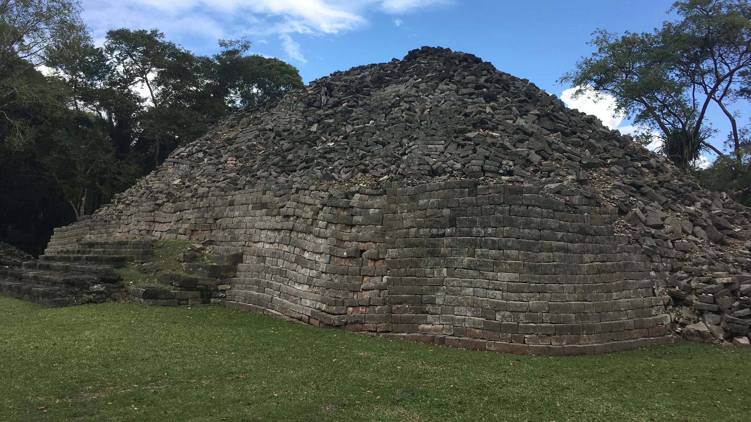 Maya stone structure with steps, surrounded by grass and trees.