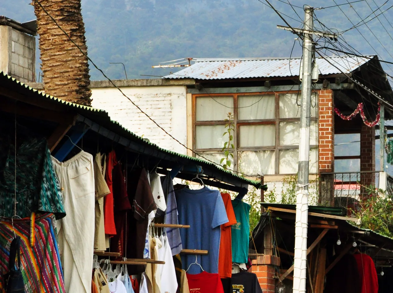 Clothes and textiles hanging at Calle Santander stalls, with buildings and mountains behind.