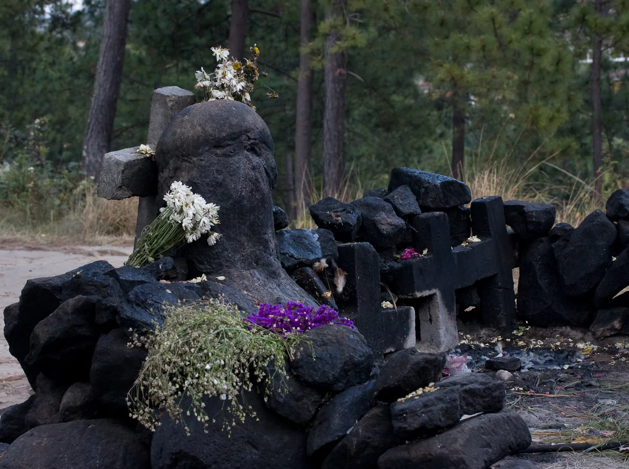 Stone and crosses at the Pascual Abaj shrine, with flowers among trees.