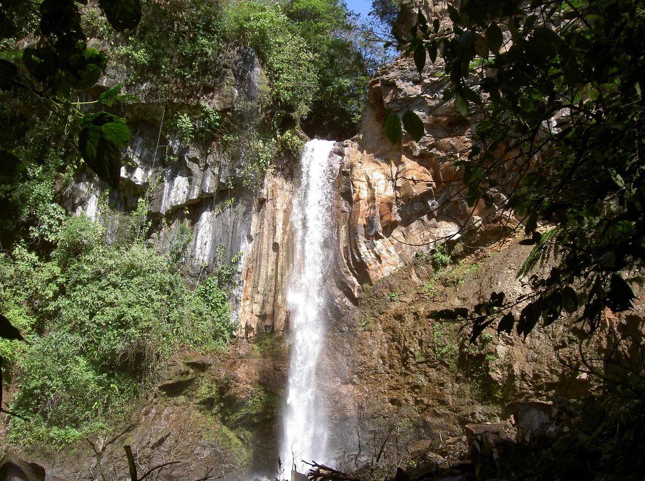 Tall waterfall between a rock face and tropical vegetation.