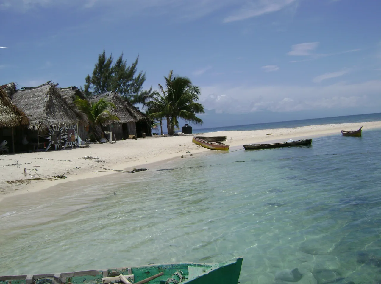Thatched houses and coconut palms on a sandy strip, with boats on clear water.