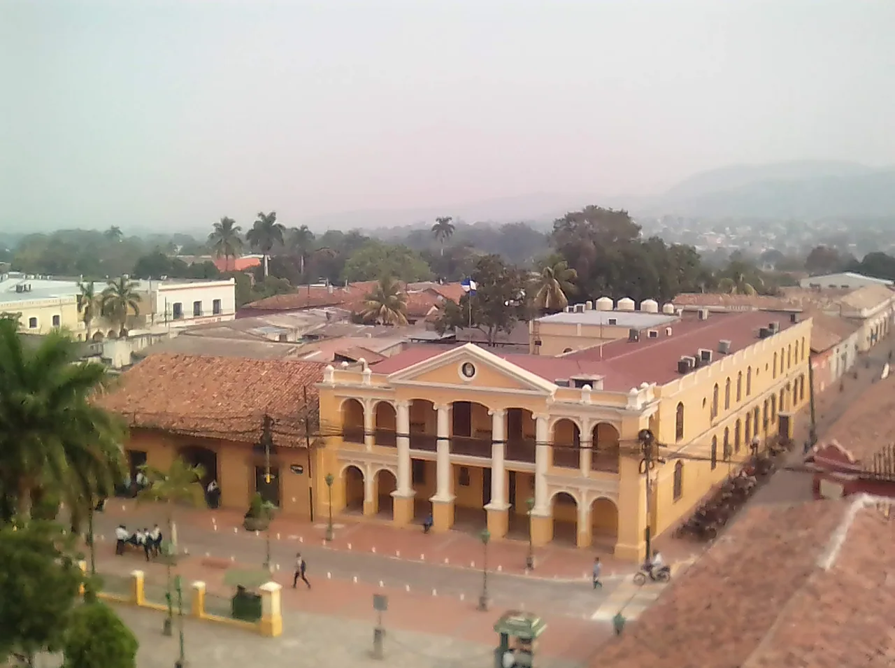 Plaza con palmeras y un palacio amarillo de arcadas, vista desde lo alto.