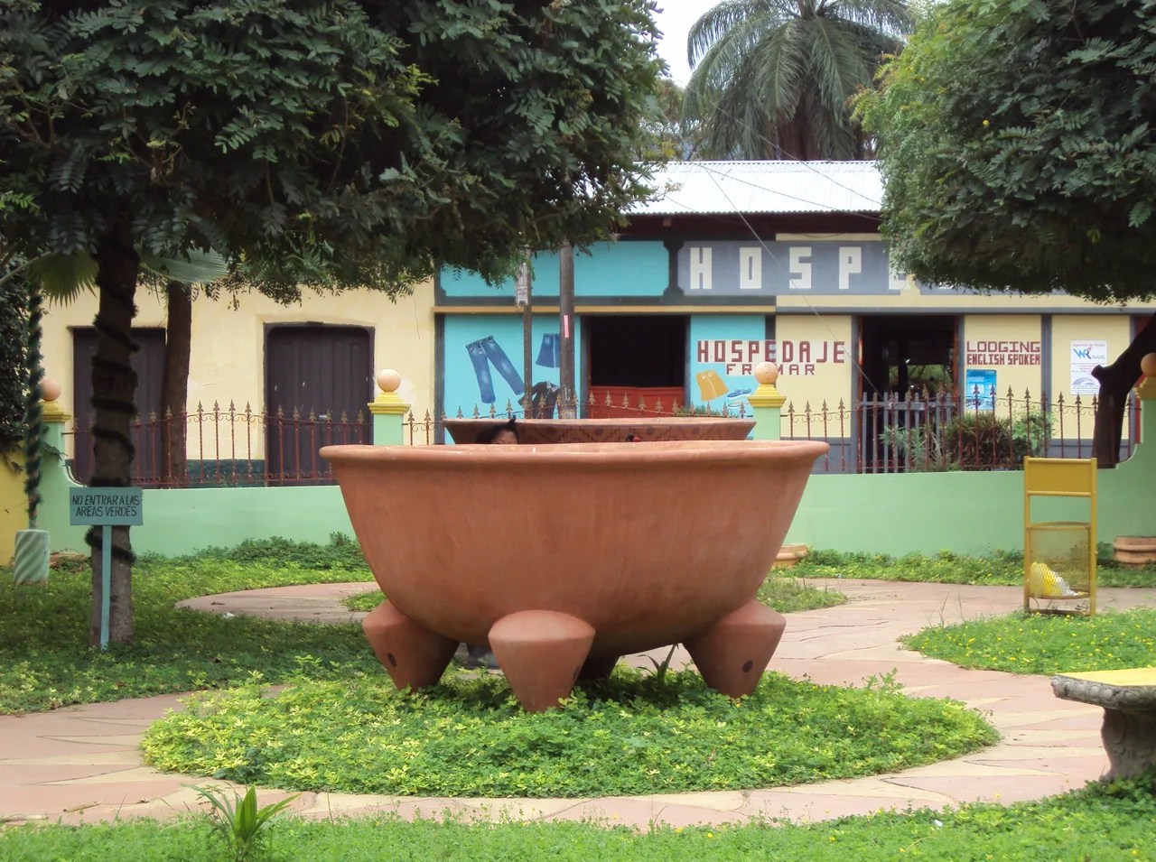 Olla grande de barro sobre tres patas en un jardín junto a casas de colores.