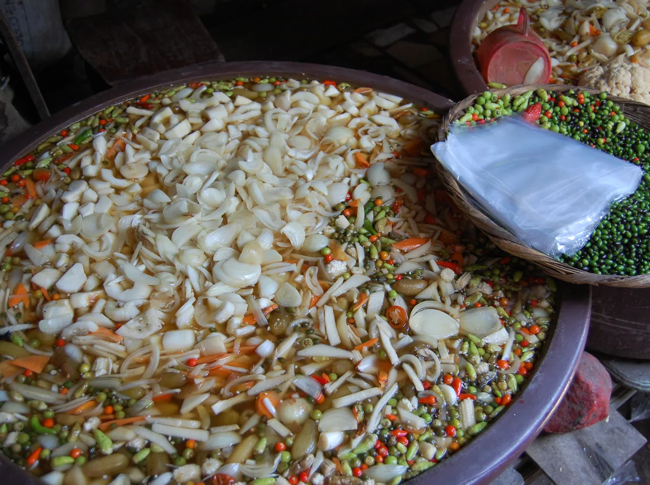 Containers of pickled onions and other vegetables beside a basket of chillies at a market stall.