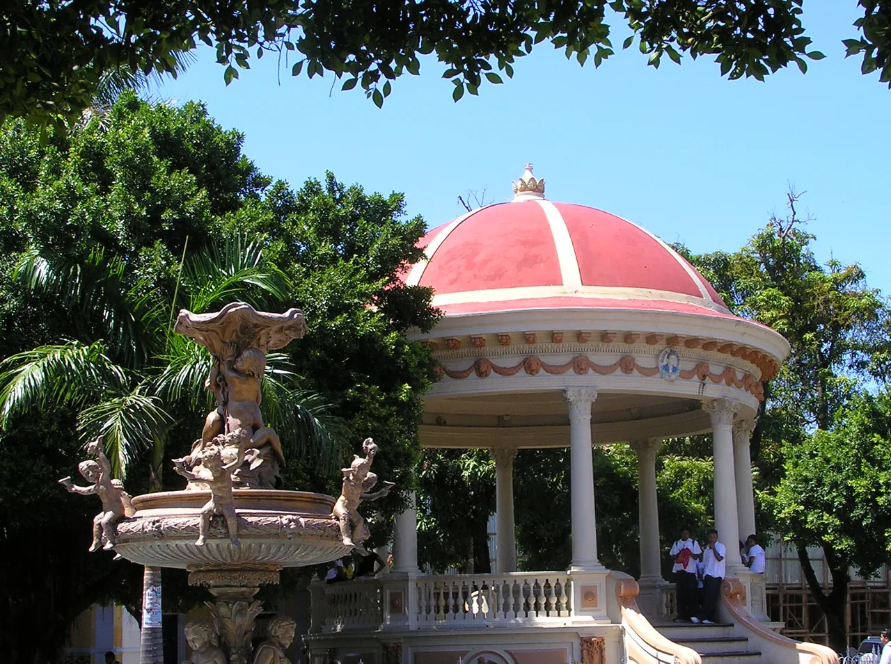 Red-roofed bandstand, fountain and lamps among park trees.
