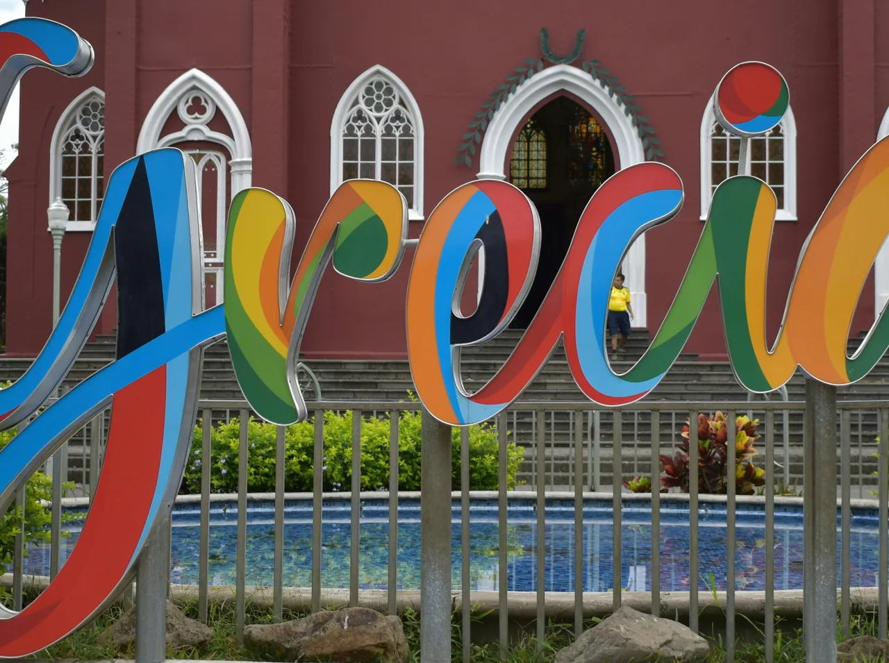 Colourful letters of the Grecia sign in front of the red church facade.