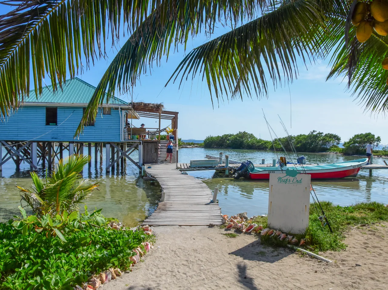 Sendero de arena entre plantas hacia un muelle y una cabaña azul sobre pilotes.
