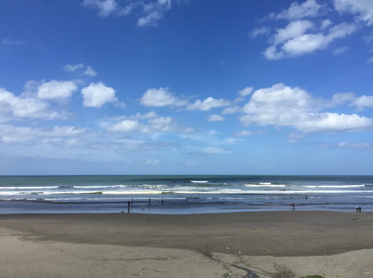 Playa ancha de arena oscura con olas blancas bajo un cielo azul con nubes.