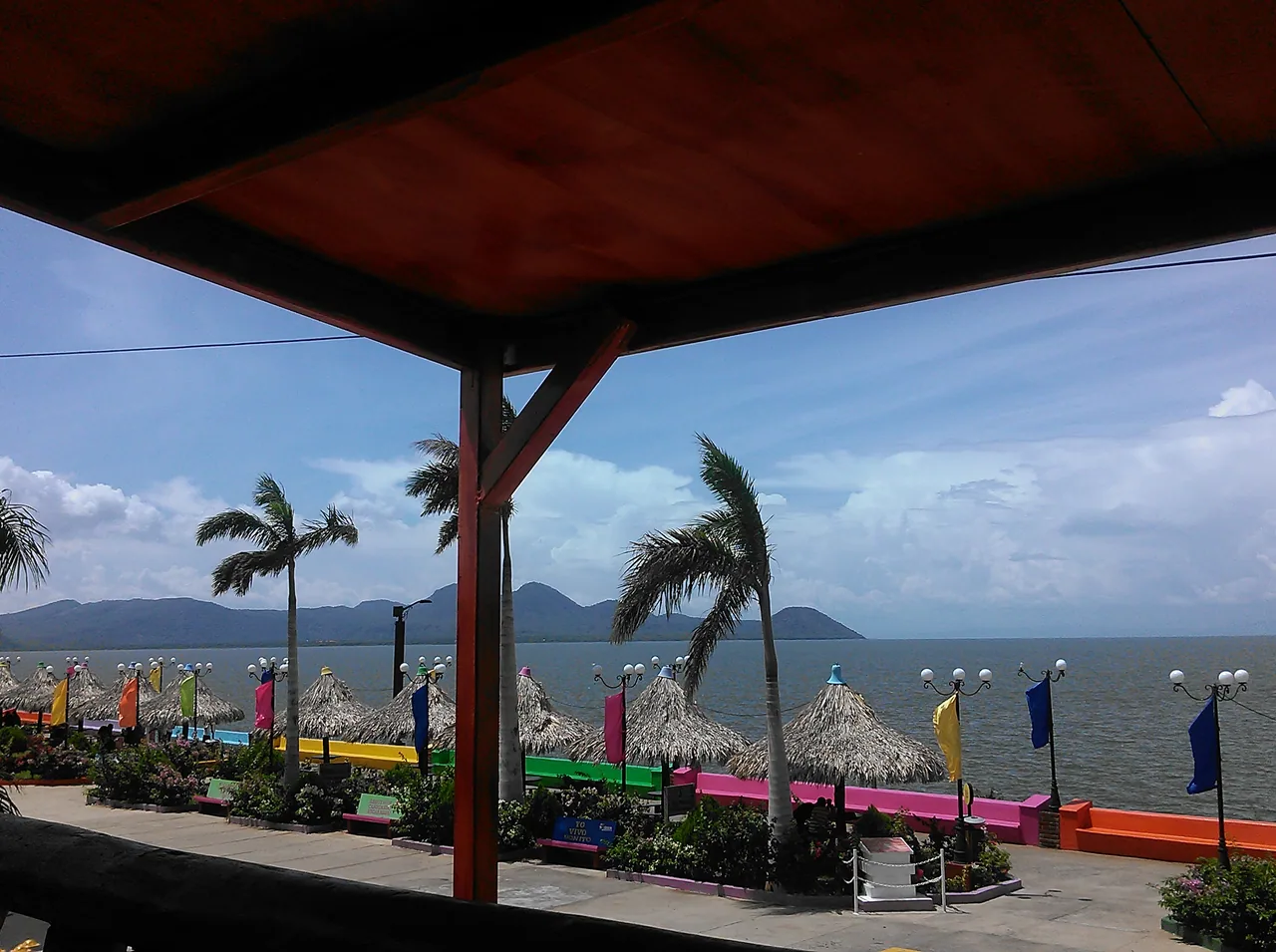 Paseo con palmeras y quioscos de colores junto al lago, visto desde una terraza.