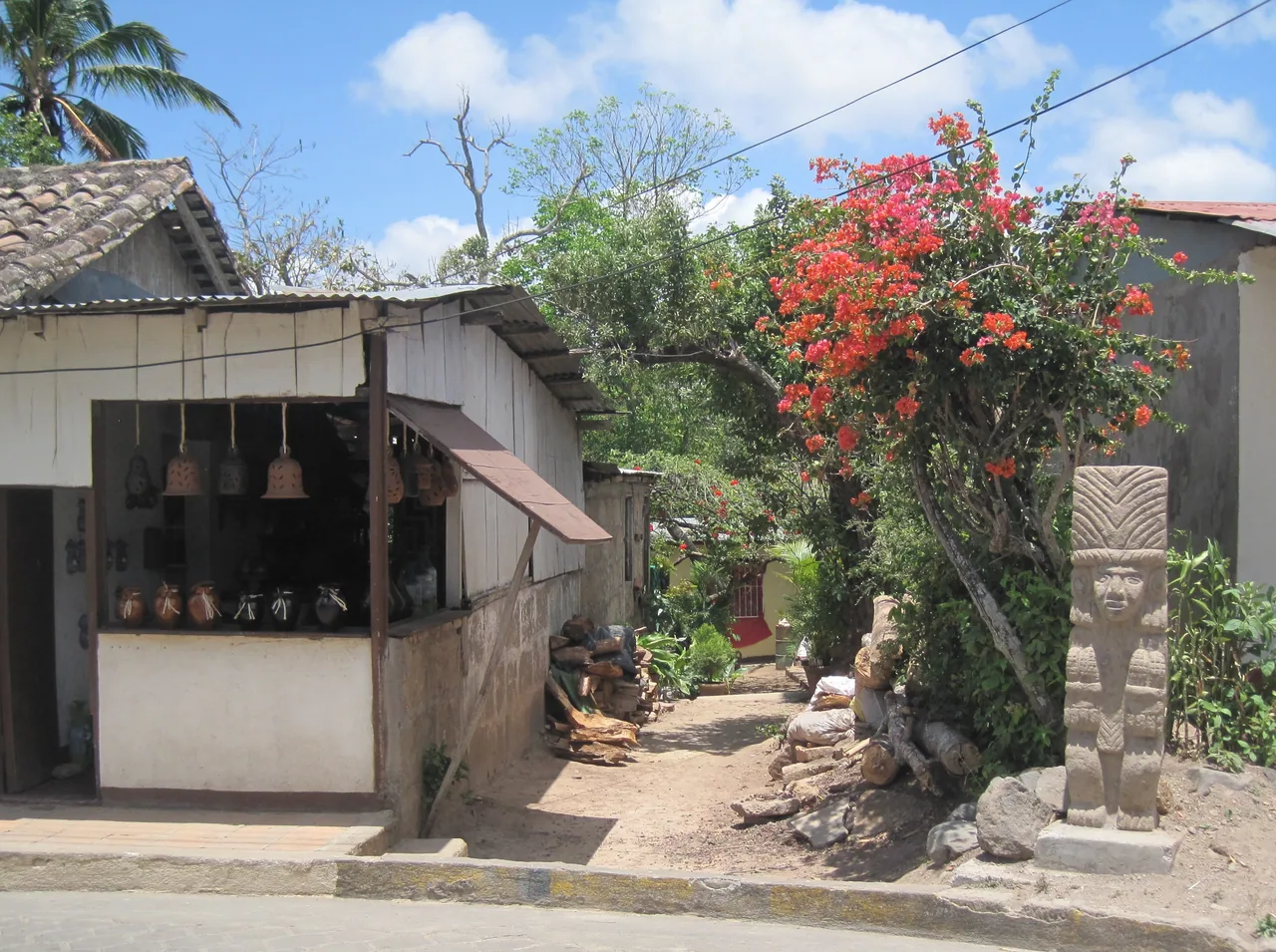 Tile-roofed workshop house with pottery, a sculpture and a red-flowering tree.