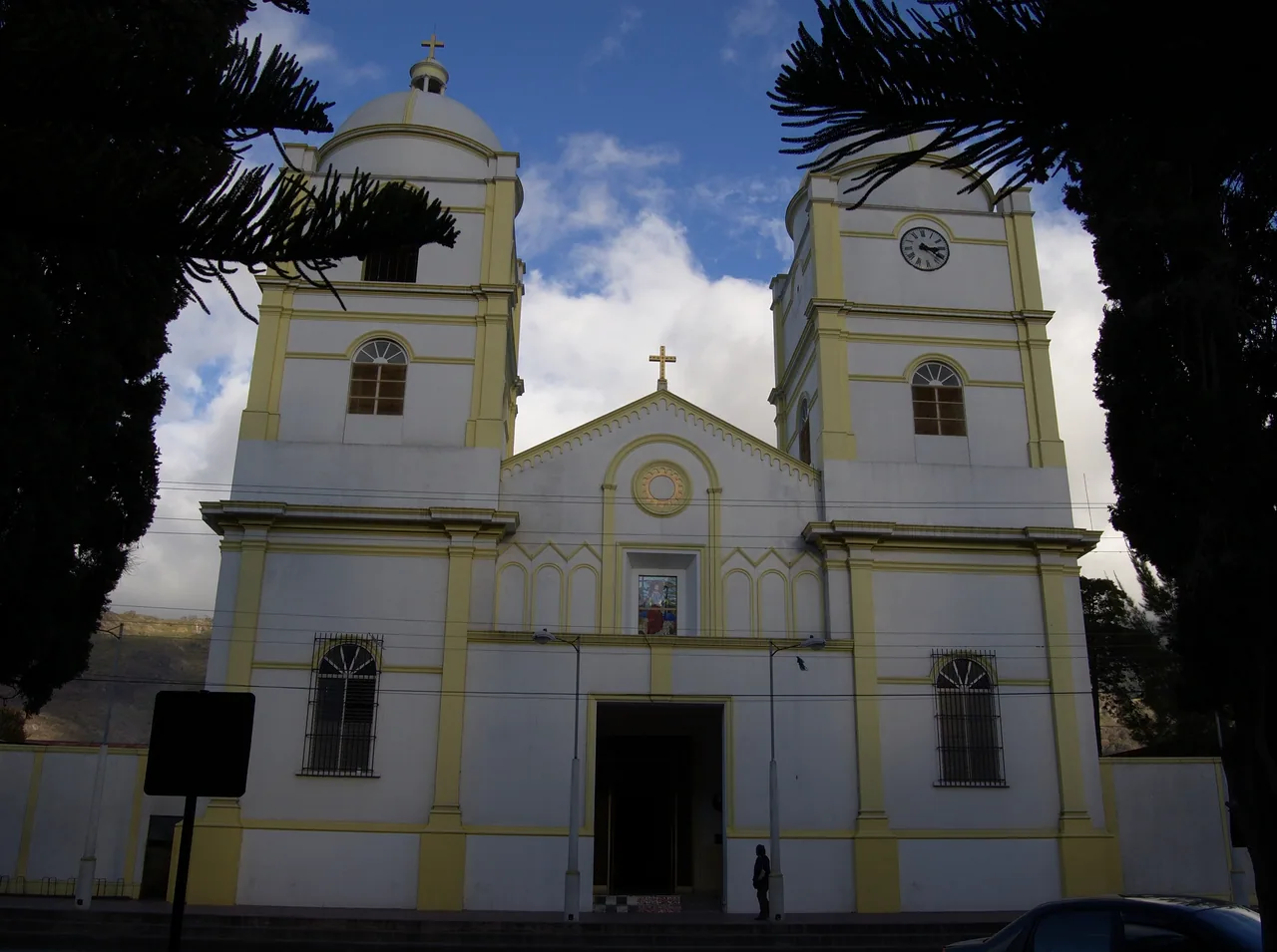 Fachada blanca y amarilla de una catedral con dos torres entre árboles.