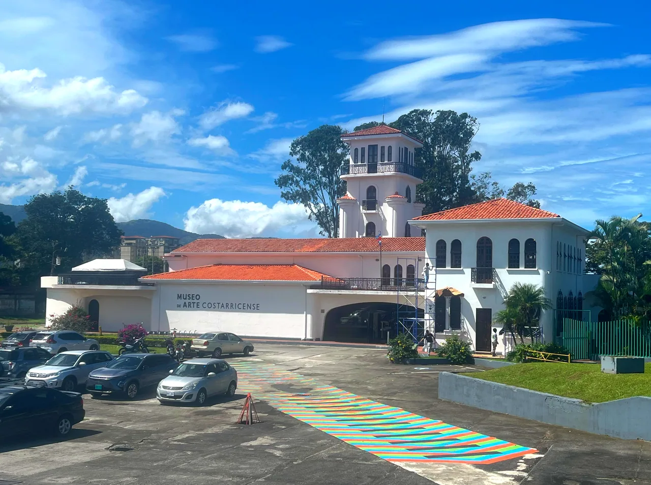 White facade and tower of the Costa Rican Art Museum, with red roofs and cars in front.