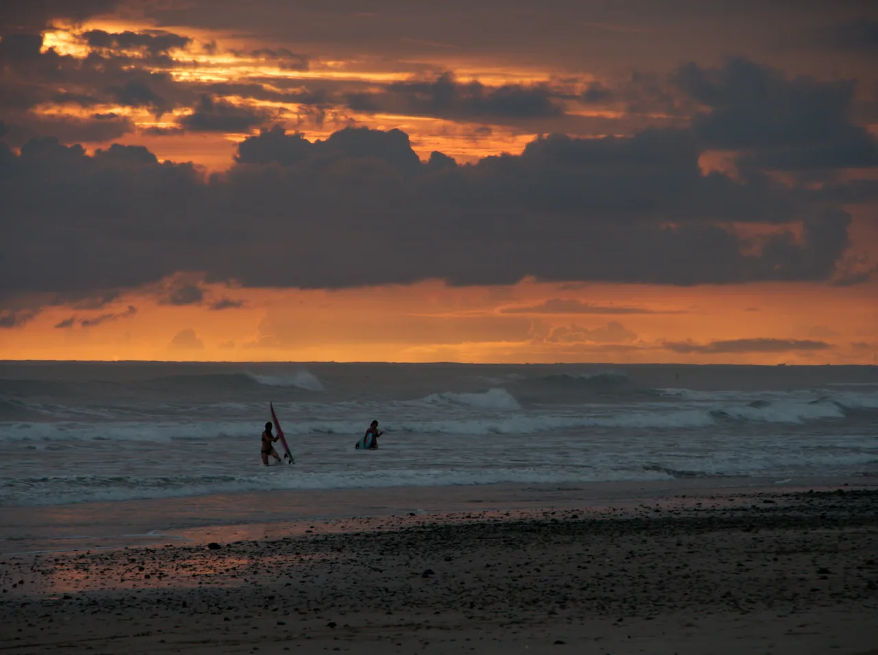 Two distant surfers entering the sea beneath an orange sunset sky at Playa Carmen.