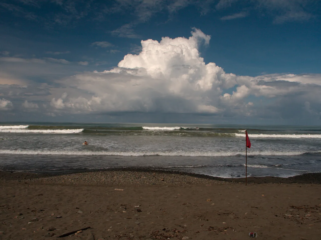 Dominical beach with waves, a red flag and large clouds over the sea.