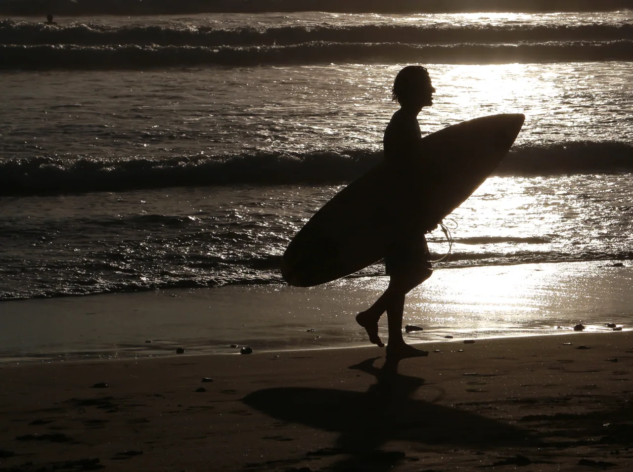 Silhouette of a surfer carrying a board beside shimmering water at sunset on Playa Hermosa.