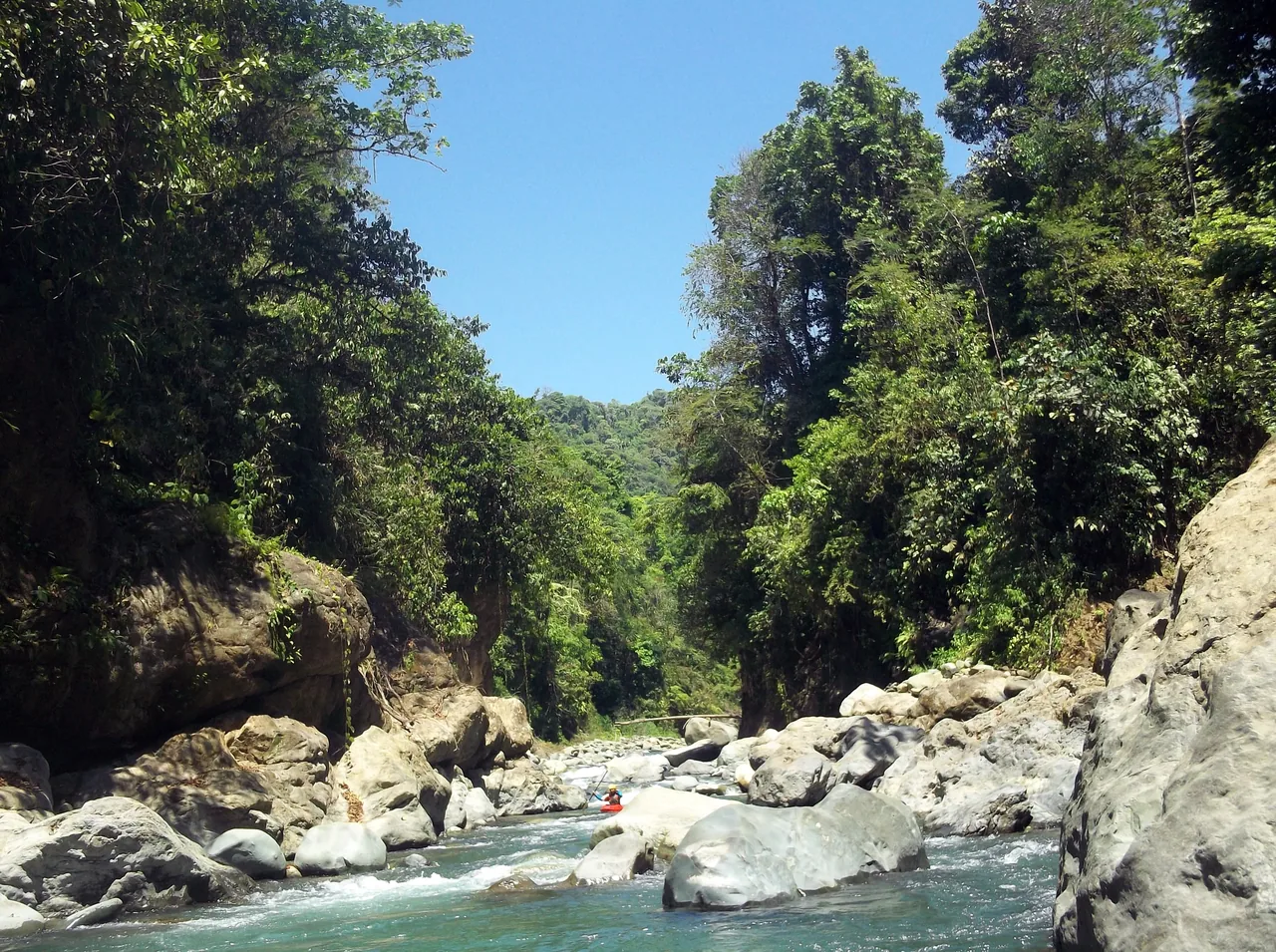The Naranjo River between large rocks and forest-covered walls, with a distant kayak.