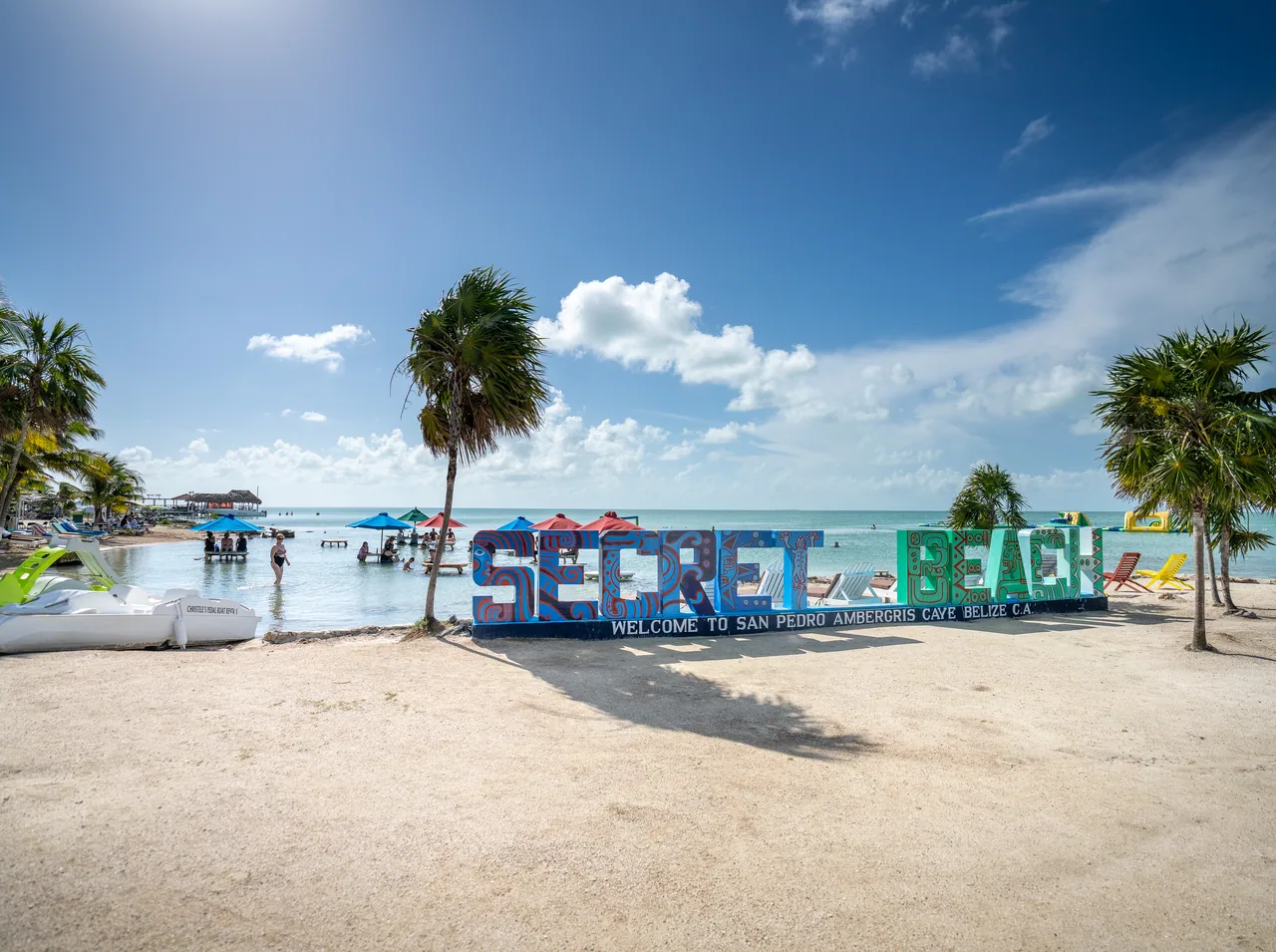 Letras de Secret Beach sobre la arena, con palmeras y agua turquesa detrás.