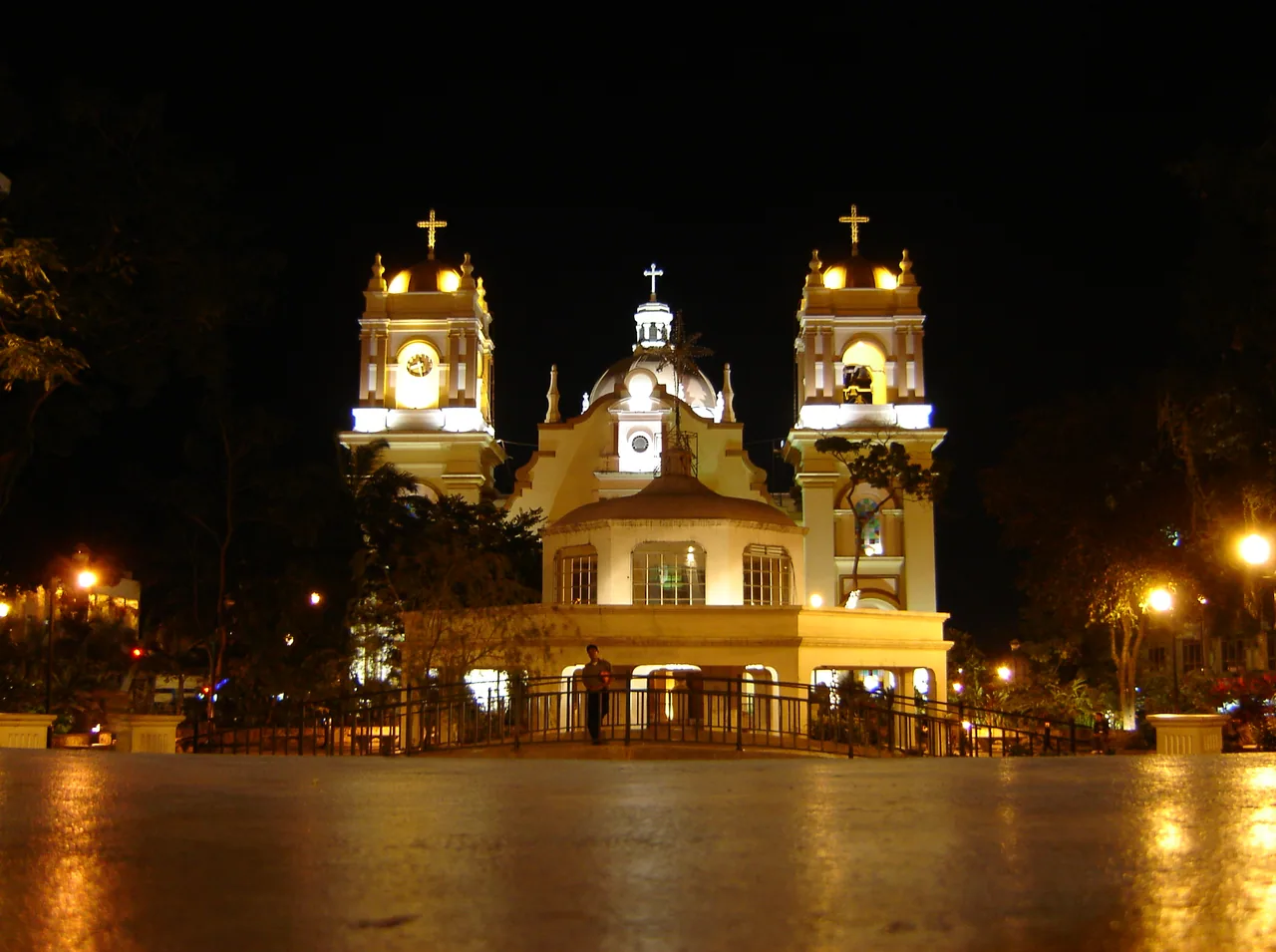 Cathedral lit at night, with two towers and a wet square in front.