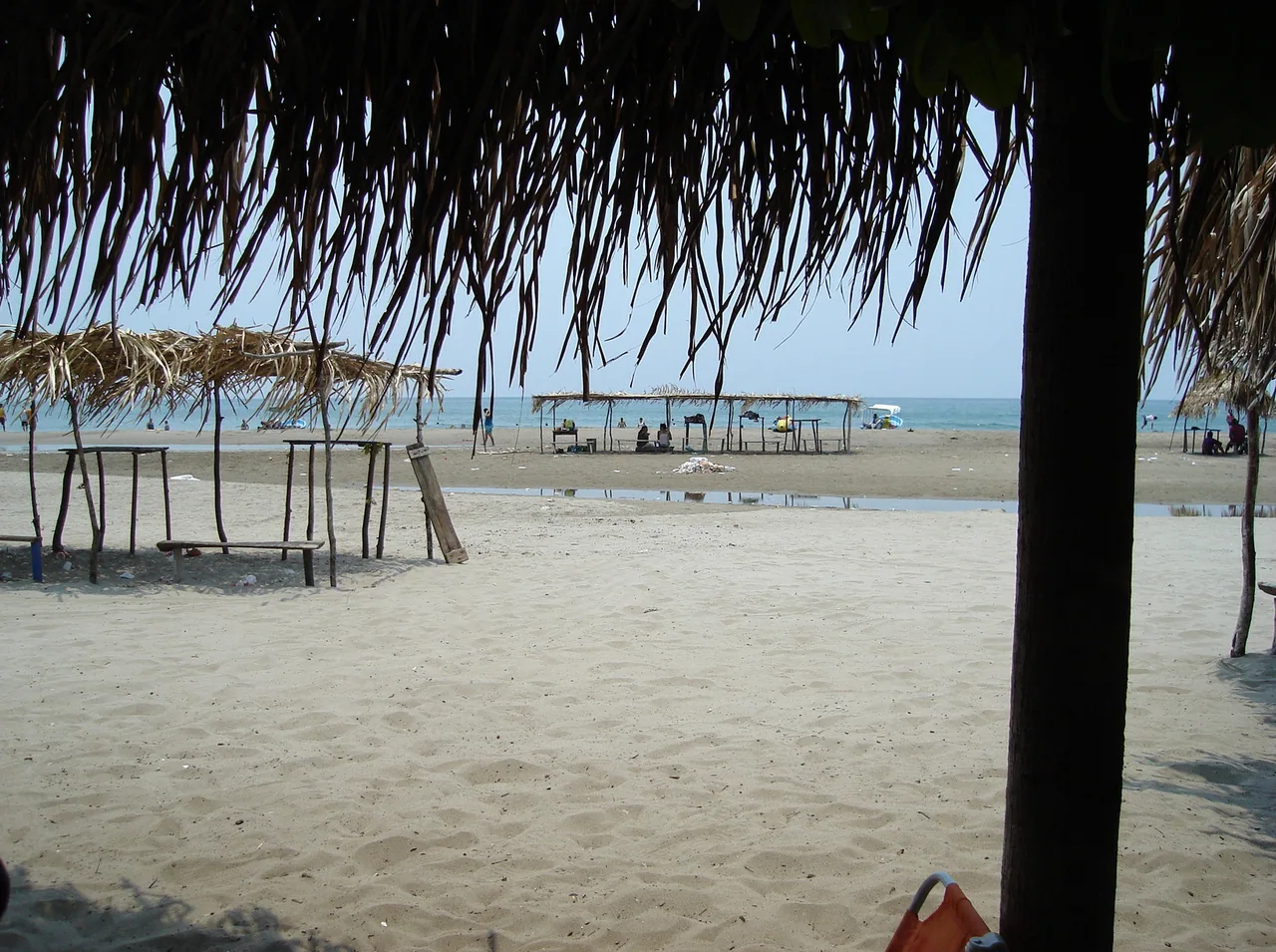 Beach with tables and palm roofs facing the sea, viewed from a thatched shelter.
